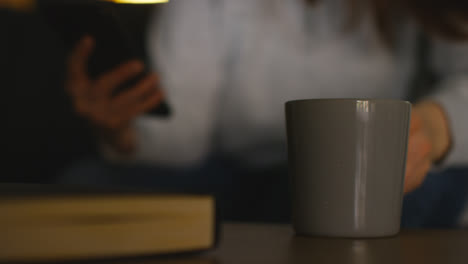 Close-Up-Of-Woman-Sitting-On-Sofa-At-Home-At-Drinking-Hot-Drink-And-Streaming-Or-Watching-Movie-Or-Show-Or-Scrolling-Internet-On-Mobile-Phone